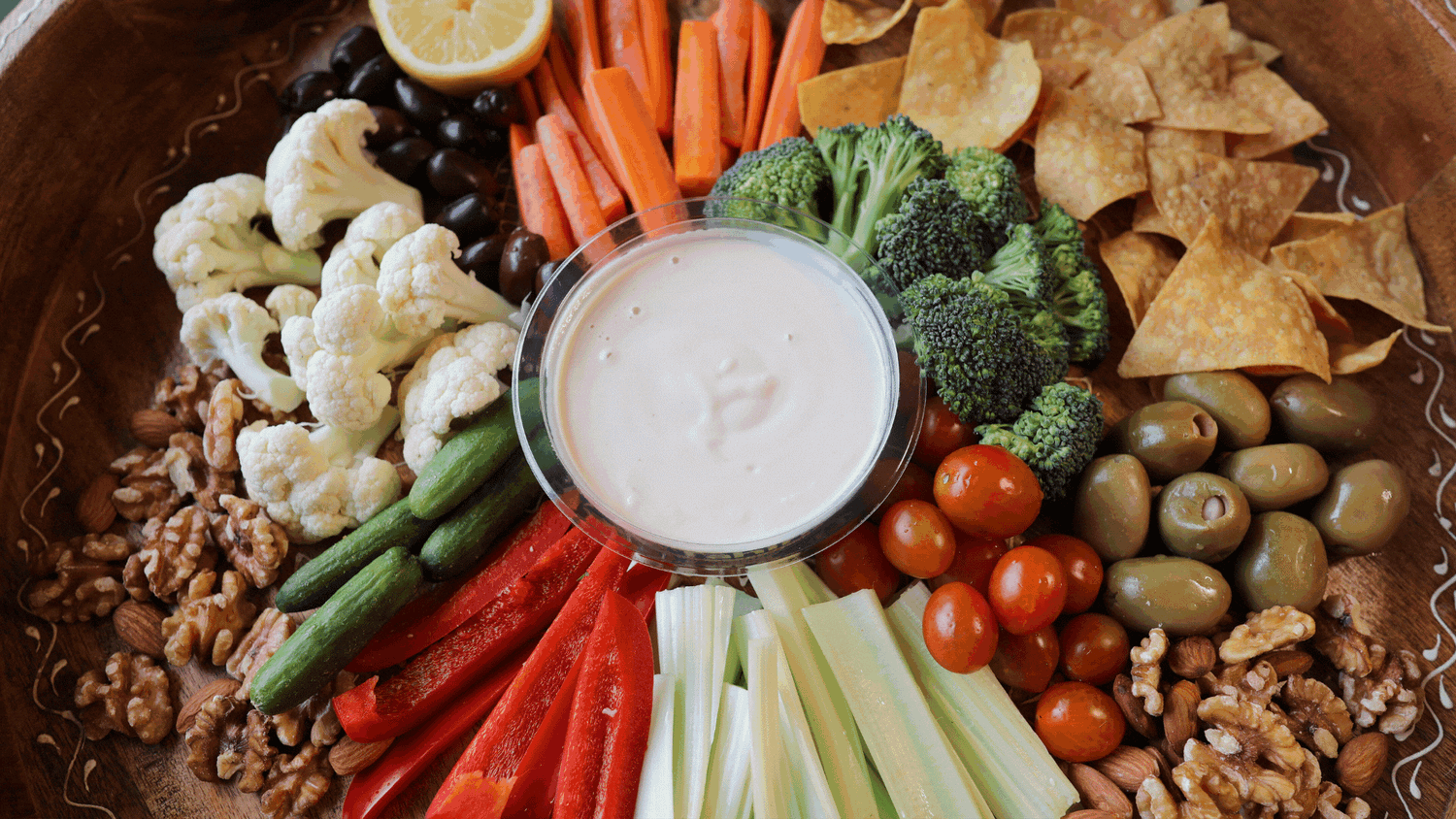 A platter featuring Tal's Mediterranean Tahini Fresh Lemon sauce surrounded by fresh vegetables, nuts, and chips for dipping. A perfect blend of creamy tahini and zesty lemon, ideal for enhancing any dish.