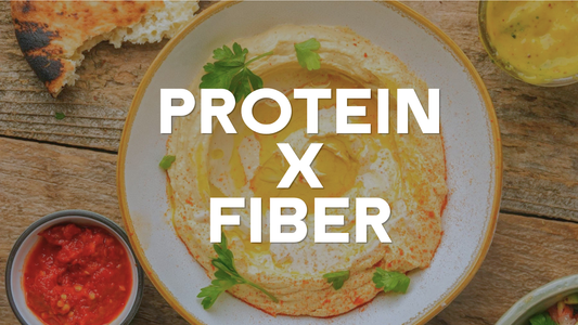 Bowl of creamy Mediterranean hummus topped with olive oil and parsley on a wooden table, surrounded by pita bread and side dishes, with the text "PROTEIN X FIBER" displayed across the image.