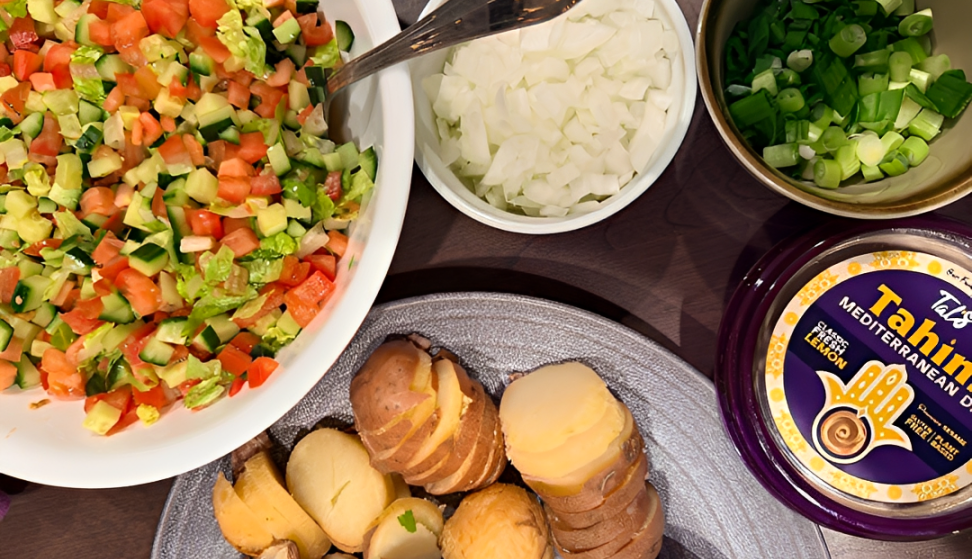 Overhead view of a Mediterranean meal featuring Tal’s Tahini Mediterranean Dip Classic Sesame Lemon alongside a chopped salad with tomatoes, cucumbers, lettuce, and carrots, a bowl of diced onions, sliced green onions, and a plate of roasted potatoes.