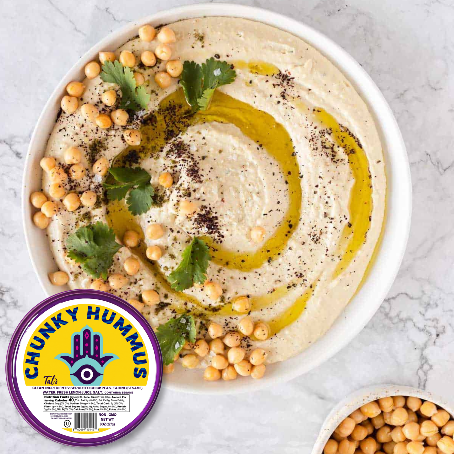 A white bowl filled with smooth hummus garnished with olive oil, whole chickpeas, fresh herb leaves, and dark seasoning, photographed from above on a light marble background. A purple and yellow Chunky Hummus Tal's hummus container appears in the bottom left corner, with additional chickpeas visible in a small bowl at the bottom right.