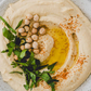 Overhead view of smooth, creamy hummus in a light-colored bowl, garnished with olive oil, whole chickpeas, fresh green herbs, and paprika seasoning. The hummus has a swirled texture and is presented as a fresh Mediterranean-style dip.