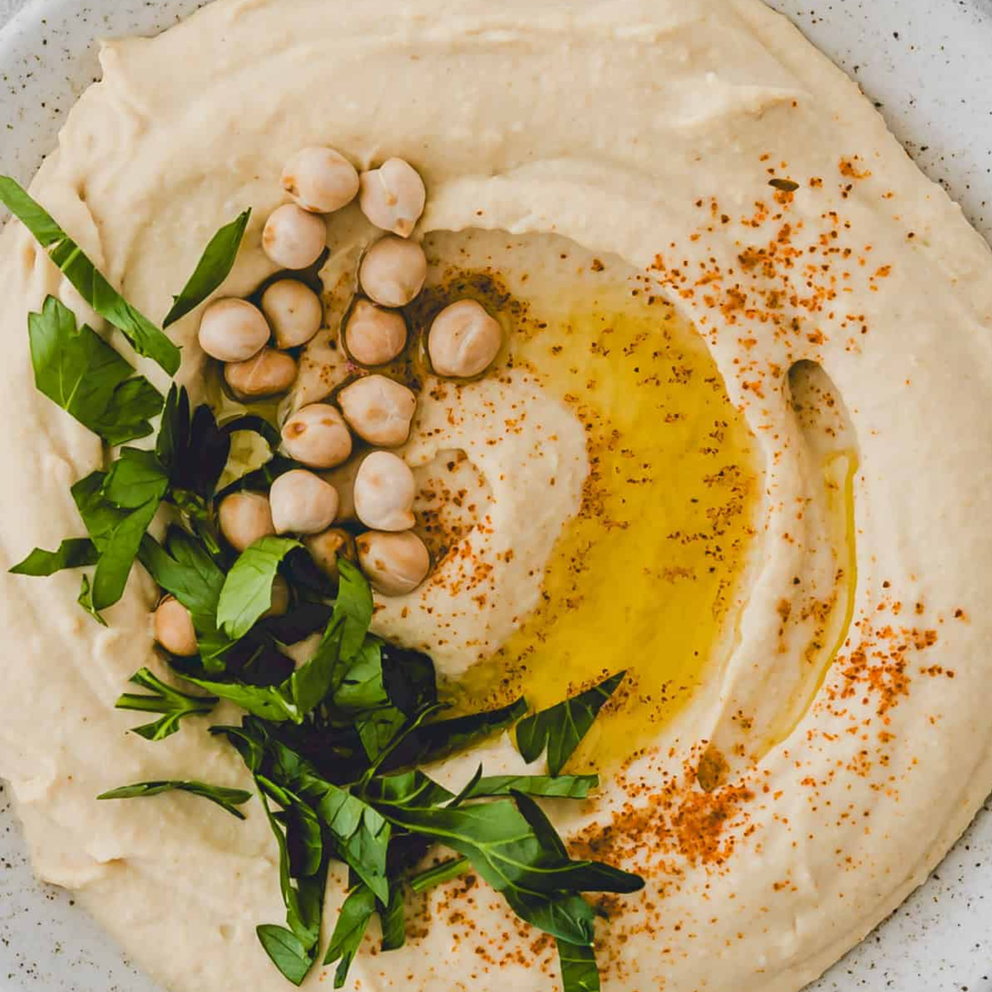 Overhead view of smooth, creamy hummus in a light-colored bowl, garnished with olive oil, whole chickpeas, fresh green herbs, and paprika seasoning. The hummus has a swirled texture and is presented as a fresh Mediterranean-style dip.