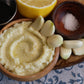 Close-up photograph of homemade Lebanese toum (garlic sauce) served in a rustic wooden bowl. The creamy, pale ivory garlic sauce has a whipped texture with a drizzle of olive oil in the center. Several peeled garlic cloves are arranged around the bowl alongside a small wooden dish of coarse salt and a lemon in the background. Natural lighting highlights the rich texture and traditional Mediterranean ingredients.