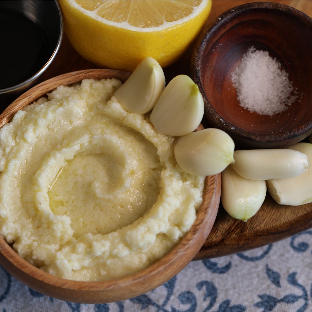 Close-up photograph of homemade Lebanese toum (garlic sauce) served in a rustic wooden bowl. The creamy, pale ivory garlic sauce has a whipped texture with a drizzle of olive oil in the center. Several peeled garlic cloves are arranged around the bowl alongside a small wooden dish of coarse salt and a lemon in the background. Natural lighting highlights the rich texture and traditional Mediterranean ingredients.